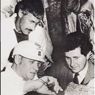 Black and white photo of three men, one wearing a helmet, looking at something together.